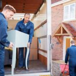 Removal Company Workers Unloading Furniture And Boxes From Truck Into New Home On Moving Day